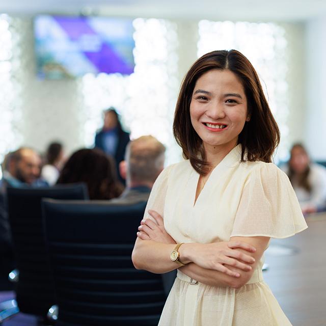 MBA student stands, smiling, while others gather around the boardroom table